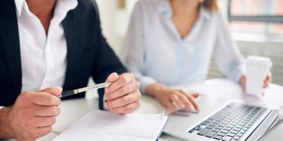 Hands of businessman with pen over open notebook during interaction with colleague